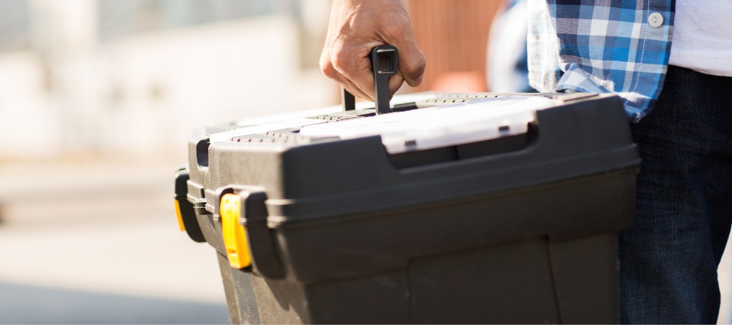 A person in a plaid shirt holds a black toolbox by the handle outdoors. The sunny setting suggests a work or repair task, conveying readiness and focus.