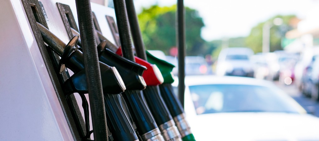 Close-up of fuel pump nozzles hanging in a gas station rack, with a blurred car and sunny street in the background; everyday travel scene.