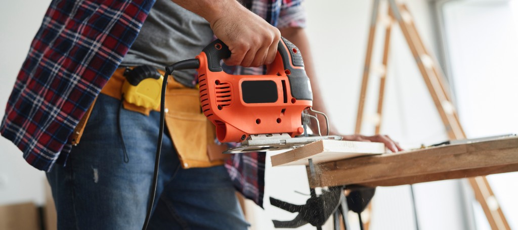 A person in a plaid shirt uses an orange jigsaw to cut wood on a workbench. A tool belt hangs around their waist. The scene conveys focus and craftsmanship.