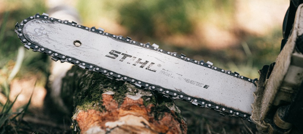 Close-up of a wet chainsaw bar resting against a freshly cut log outdoors, with water droplets, blurred greenery, and a rugged mood.