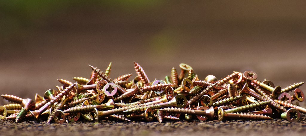 A close-up of a pile of golden screws scattered on a rough surface. The screws are shiny, with visible thread detail, set against a blurred brown background.