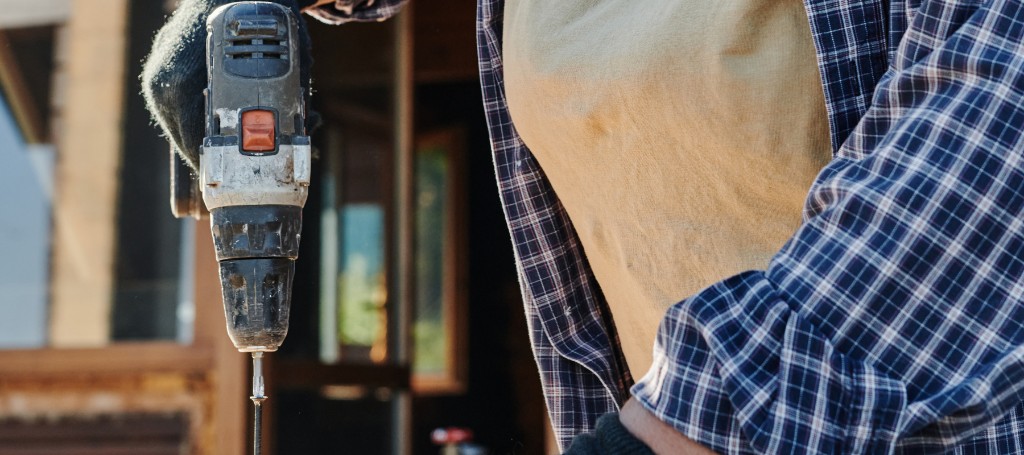 Worker wearing a plaid shirt and gloves, holding a power drill. The focus is on the drill, suggesting construction or DIY work, with a sunny outdoor setting.