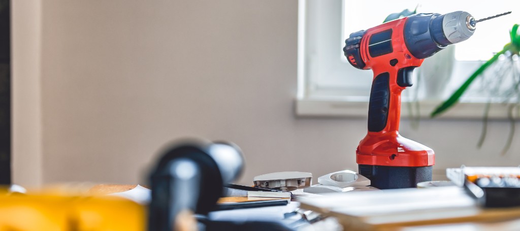 A red cordless drill stands on a cluttered wooden table near a window. Sunlight filters in, casting a warm glow. The scene conveys a sense of home improvement.