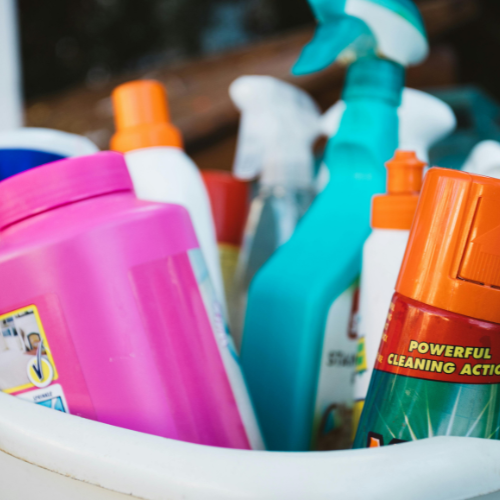 Close-up of a variety of colorful cleaning product bottles in a container, featuring spray bottles and detergent containers, suggesting cleanliness and tidiness.