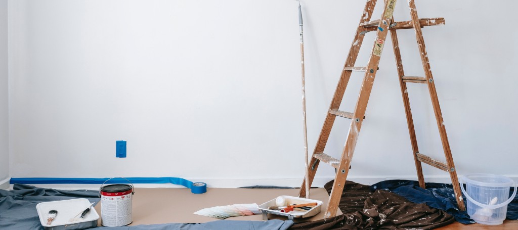A bright room mid-renovation, with a wooden step ladder, paint cans, brushes, rollers, drop cloths, and blue painter's tape.