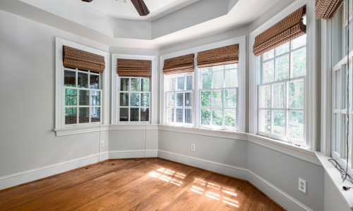 Sunlit empty room with hardwood floors and a bay window. Natural light streams through six windows with brown woven shades, creating a warm, serene atmosphere.
