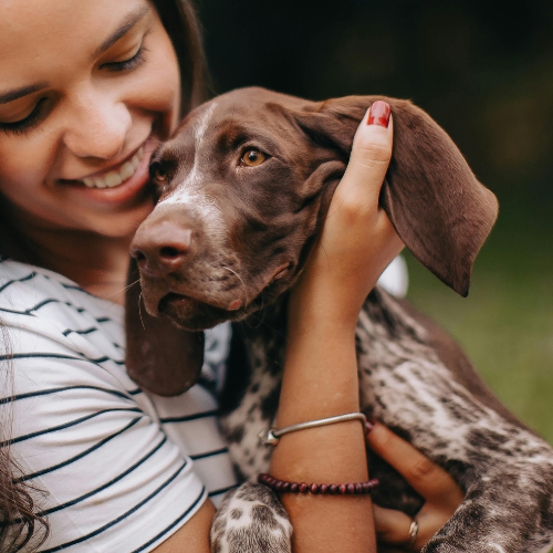Woman smiling while holding a brown puppy with floppy ears. She wears striped shirt and bracelets. The scene conveys warmth and affection outdoors.