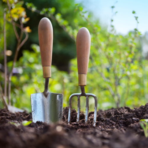 Two wooden-handled gardening tools, a trowel and a fork, are embedded upright in rich soil. Lush green plants and a soft-focus background evoke a serene garden scene.