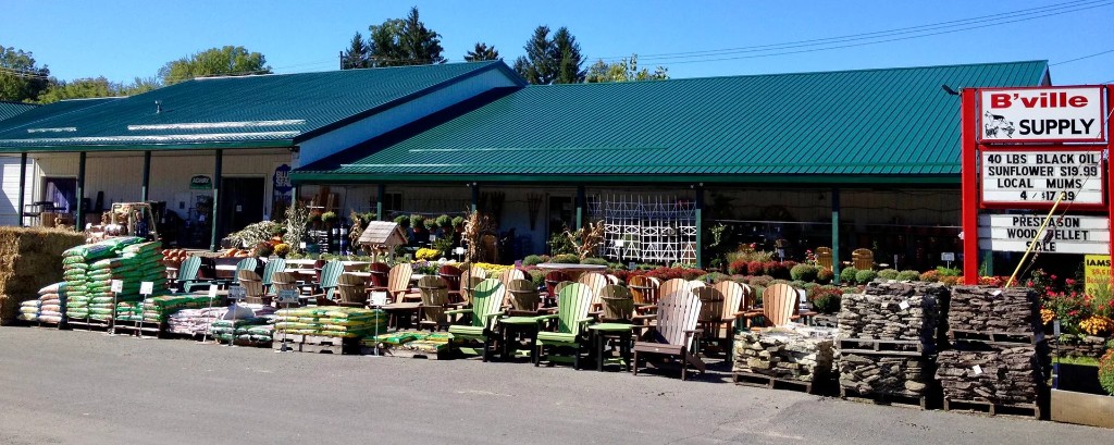 B'ville Supply garden center displays outdoor furniture, plants, landscaping stones, and gardening supplies outside a green-roofed store.