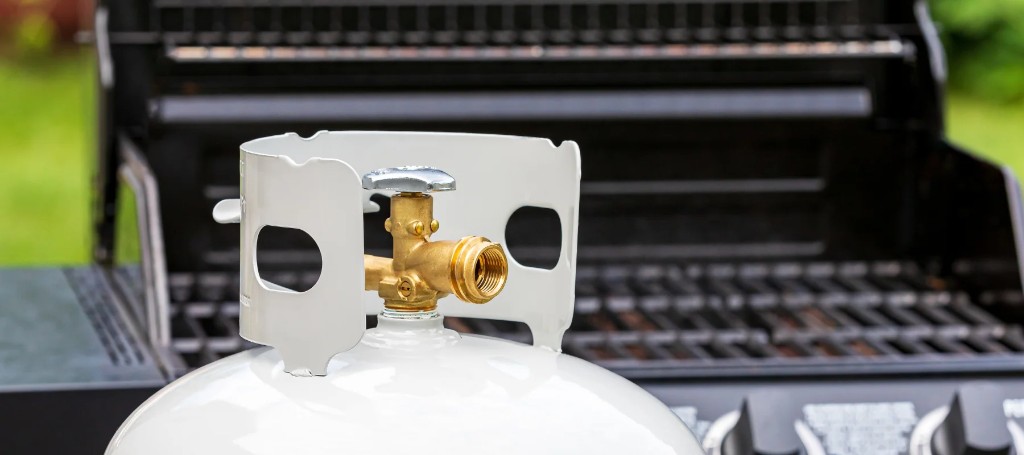 A white propane tank with a brass valve sits in front of an open black barbecue grill, suggesting a ready-to-use outdoor cookout.