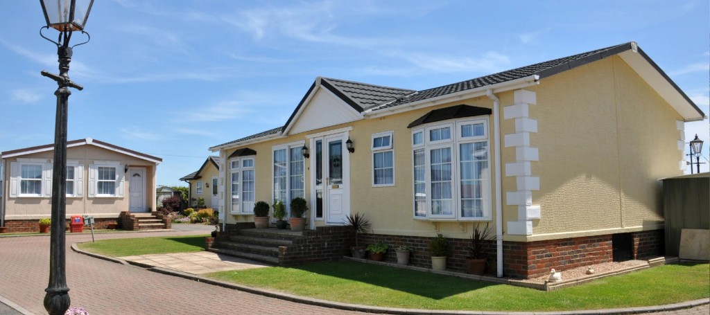 Sunny residential neighborhood with a yellow-and-white bungalow, manicured lawns, brick walkway, vintage streetlamp, and blue sky.