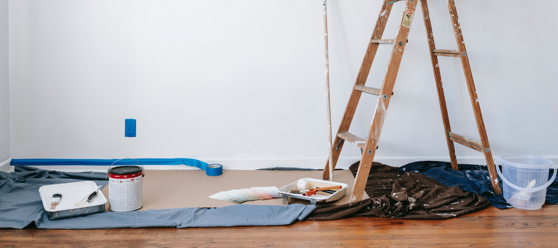 Home renovation in progress: wooden ladder, paint cans, brushes, trays, and protective tarps sit before a freshly painted white wall.