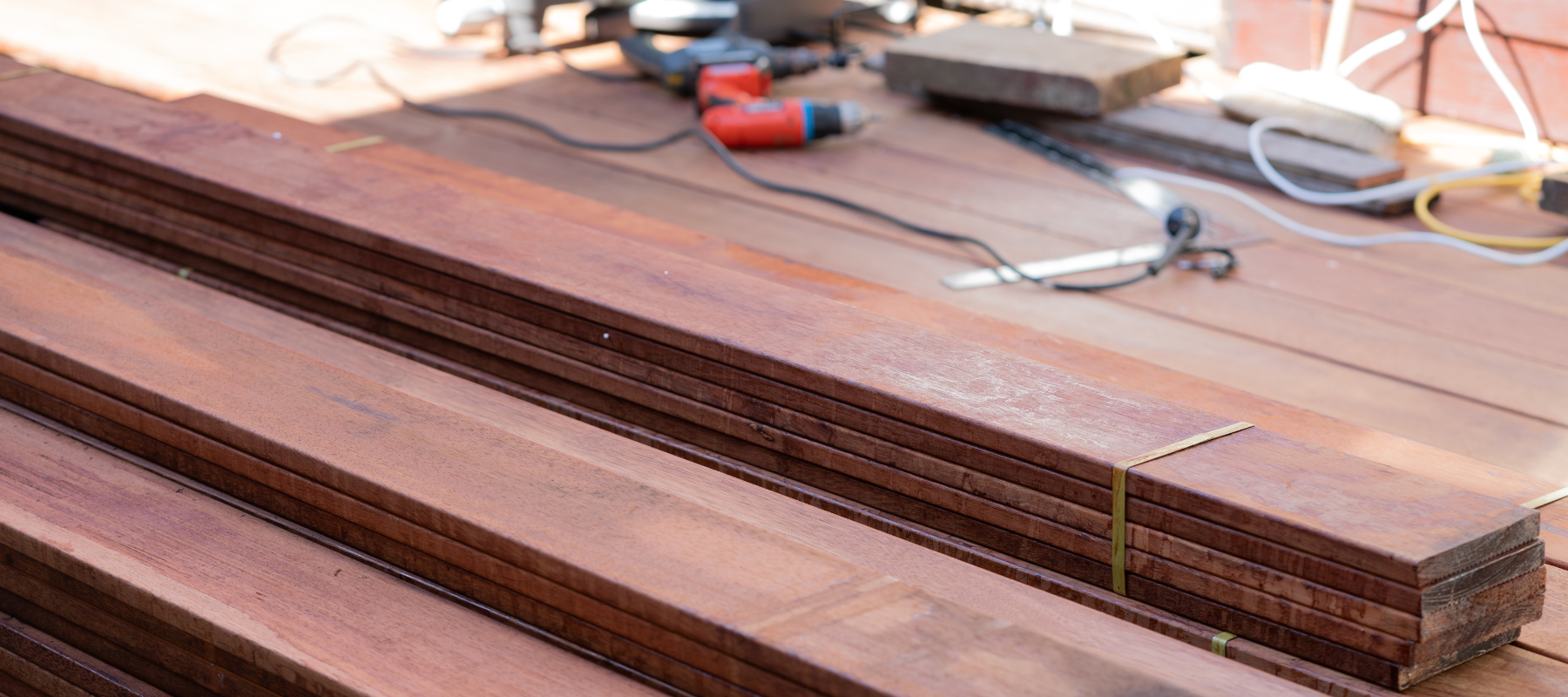 Stacked dark-stained wooden boards rest on a workbench, with power tools, cords, and hardware in the background, suggesting woodworking.