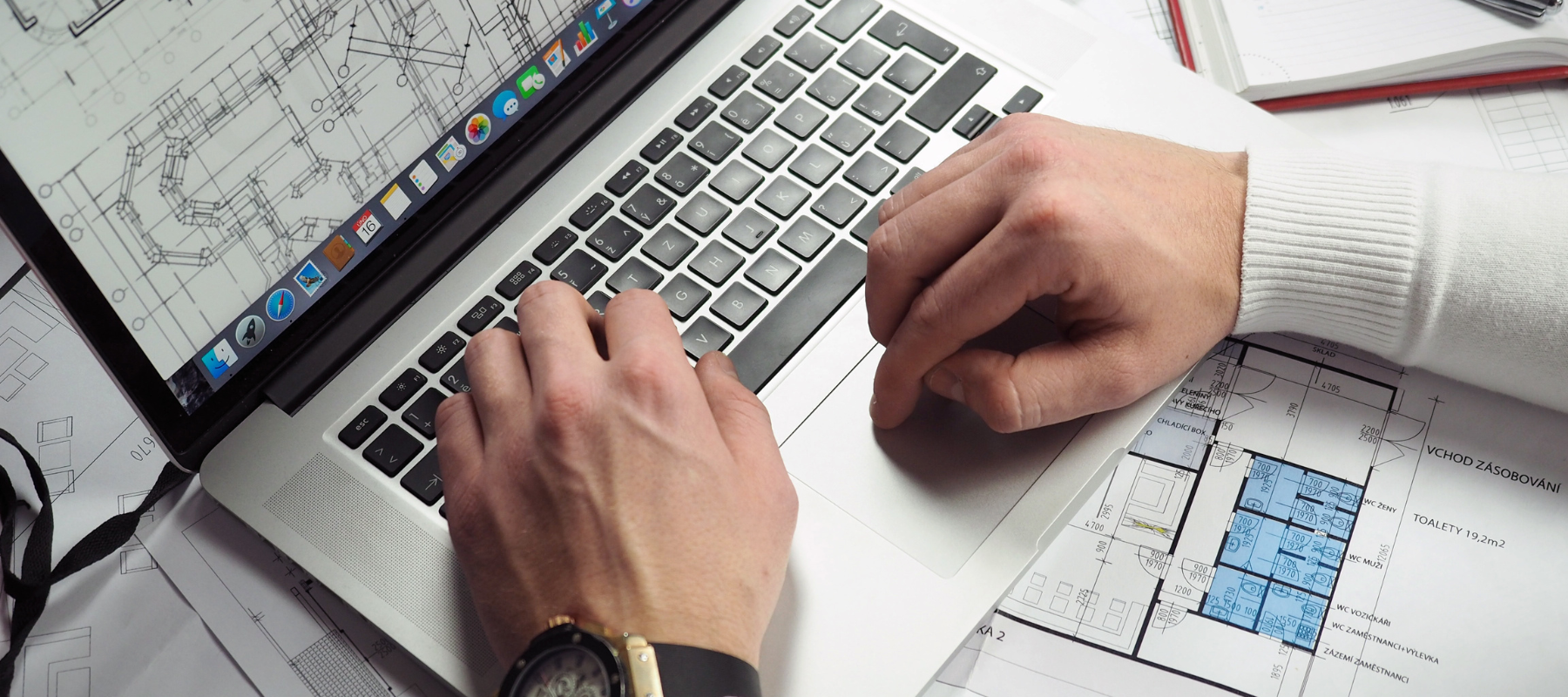 Architect using a laptop over architectural blueprints, with floor plans and a highlighted blue room visible; focused, professional mood.