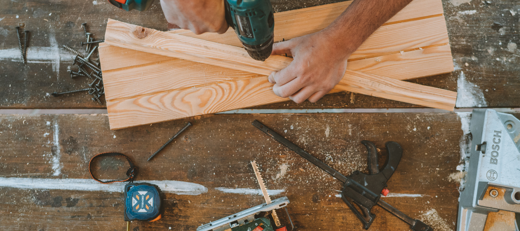 Hands use a cordless drill to fasten a wooden board on a workbench, surrounded by clamps, tools, sawdust, and measuring equipment.
