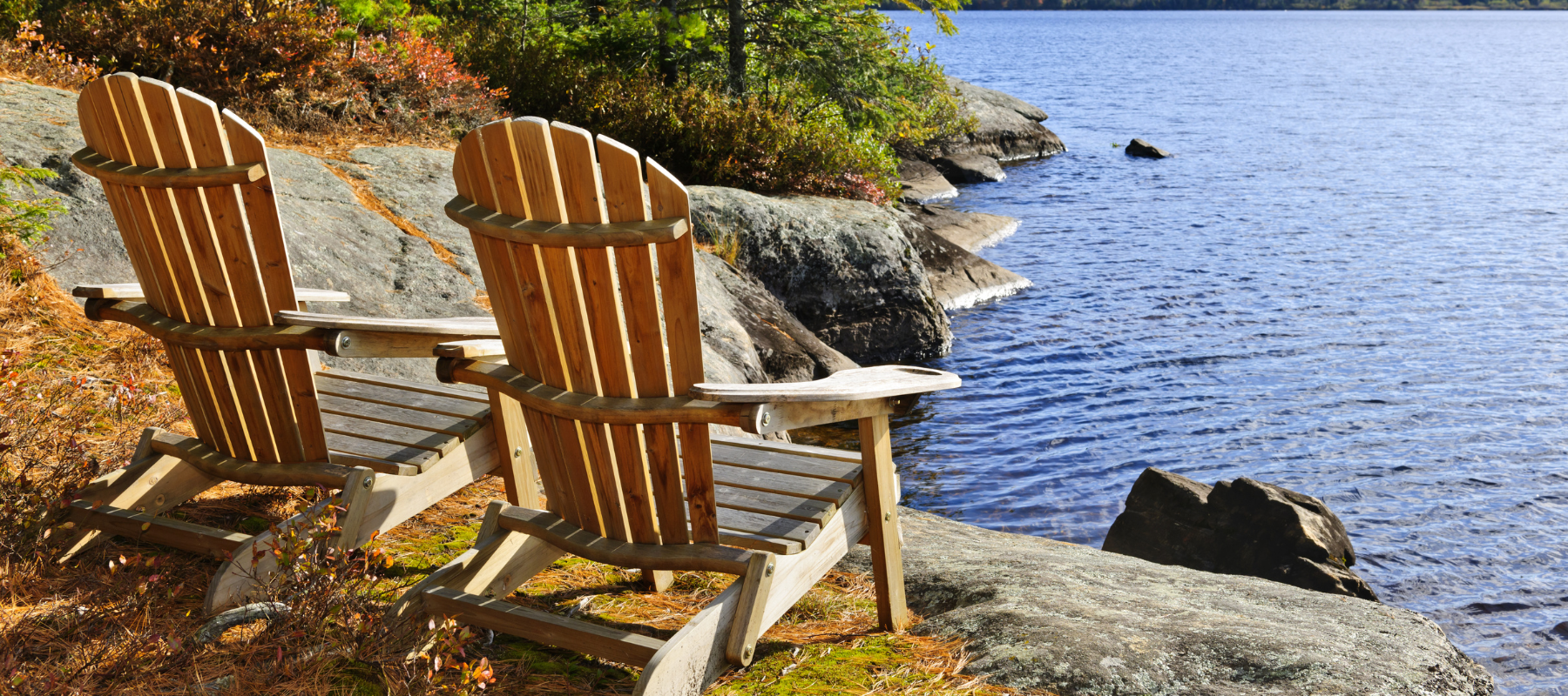 Two wooden Adirondack chairs overlook a calm blue lake from a rocky shore, framed by autumn foliage in a peaceful outdoor setting.