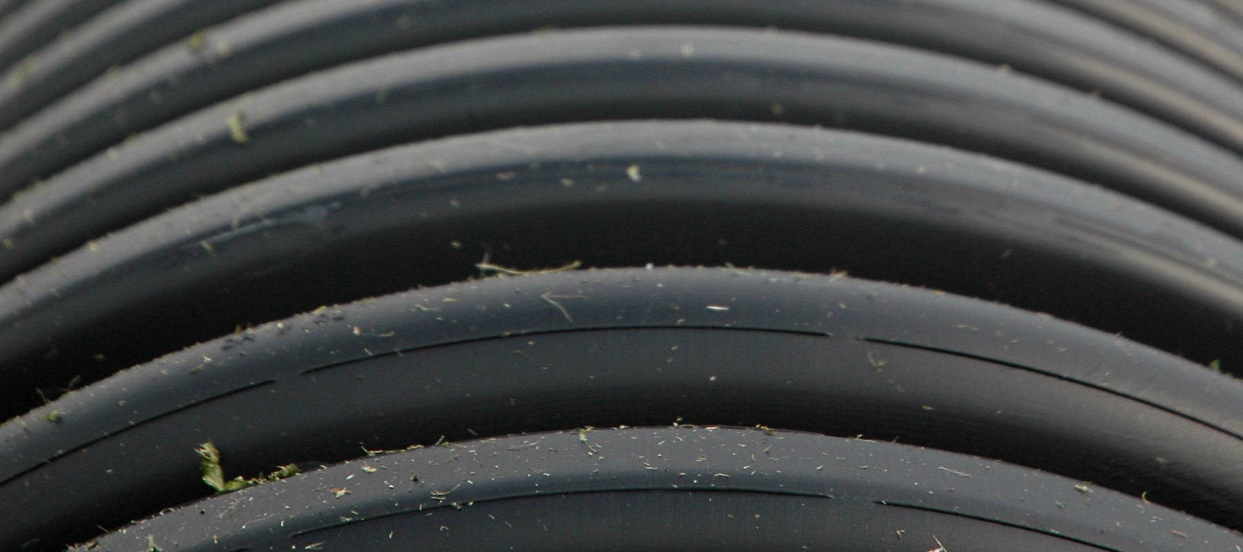 Close-up of tightly coiled black hoses or tubing, layered in curved bands with scattered dust, grass, and debris, creating an industrial texture.