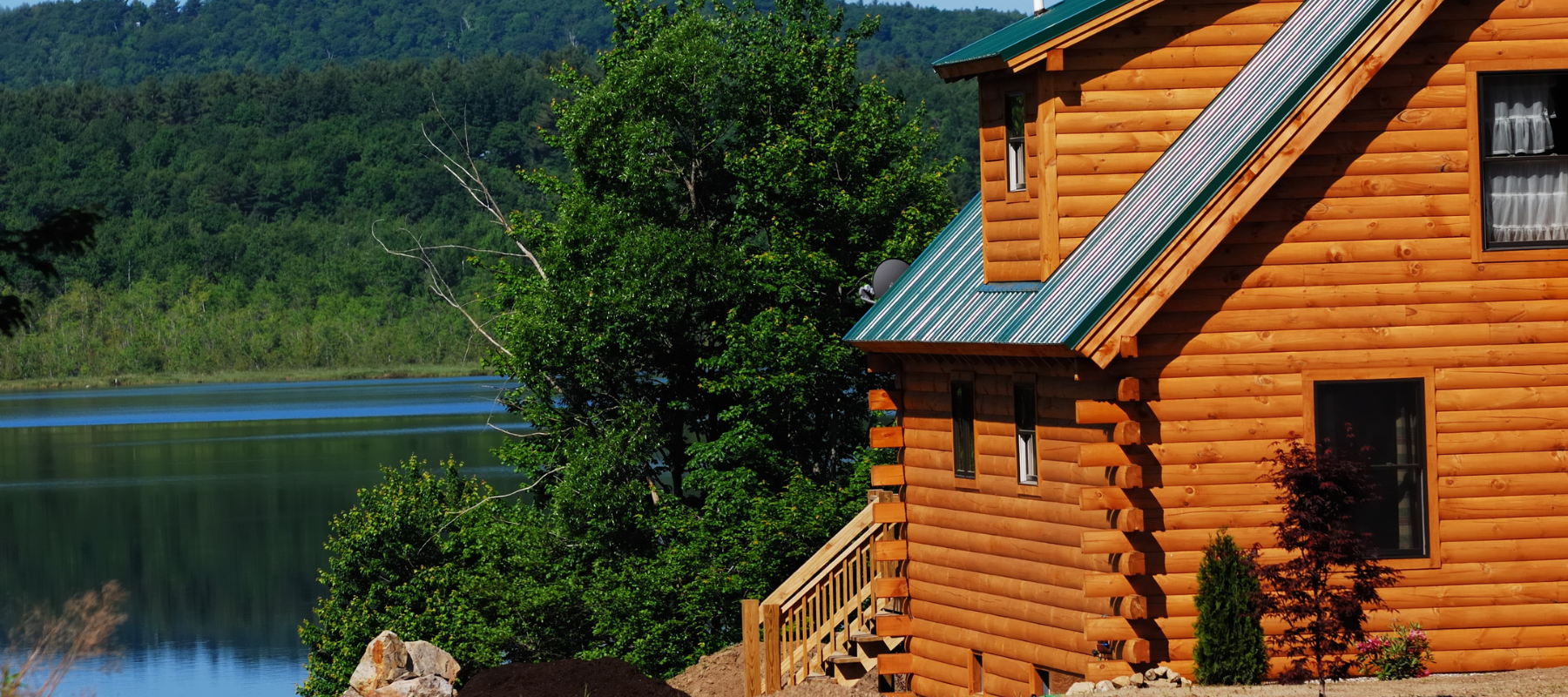 Rustic log cabin with a green metal roof overlooks a calm lake, surrounded by lush forest and mountains, creating a peaceful, secluded retreat.