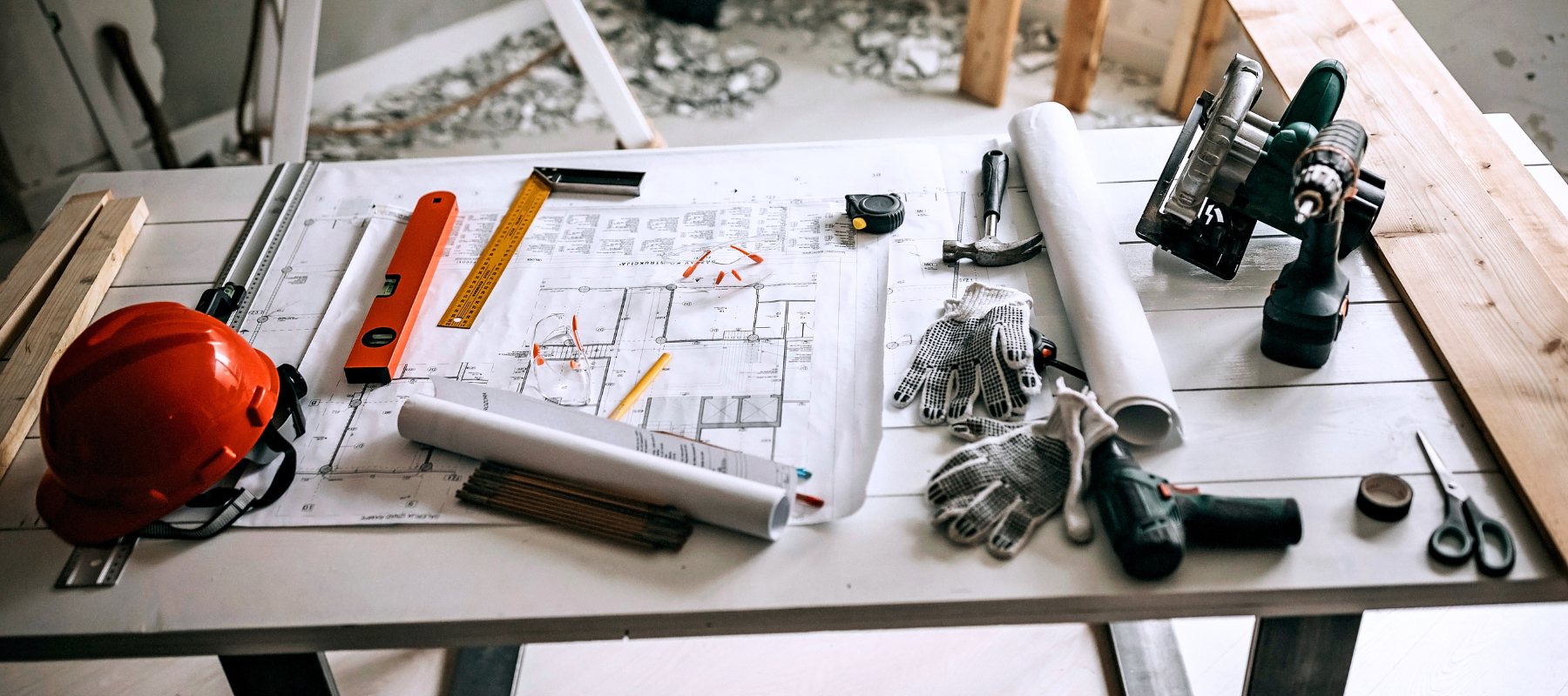 Architectural blueprints, red hard hat, tools, safety gloves, and measuring equipment cover a workshop table, conveying active construction planning.