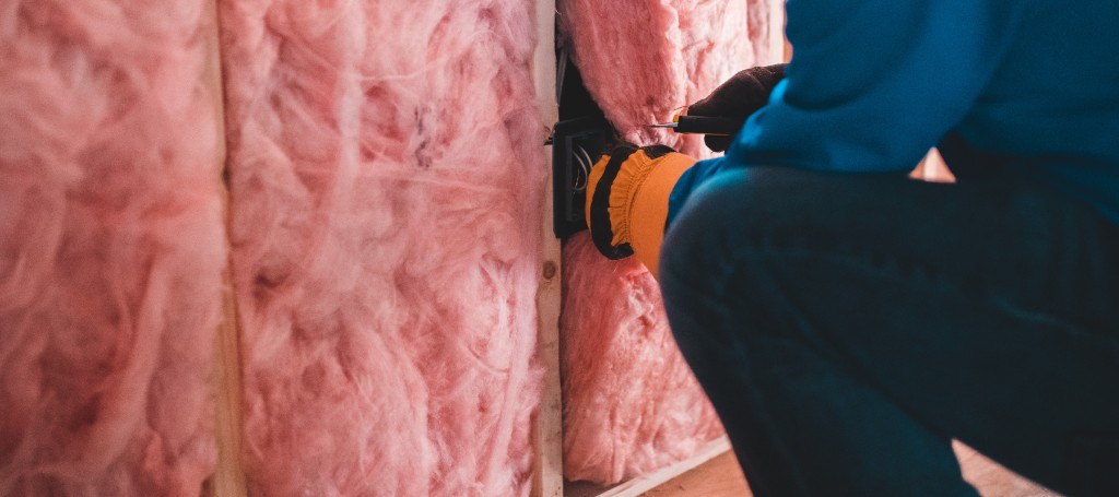 A person in a blue jacket and gloves installs pink fiberglass insulation in a wooden frame, emphasizing warmth and home construction.