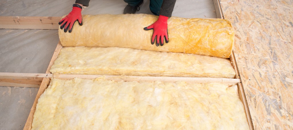 A person wearing red gloves is installing yellow fiberglass insulation between wooden floor joists. The scene conveys a sense of careful, manual labor.