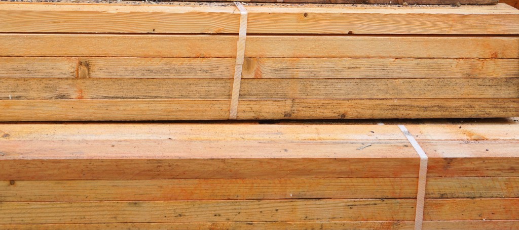 Stacks of light brown wooden planks, neatly aligned and secured with straps, suggesting a construction or woodworking context. The tone is practical and orderly.