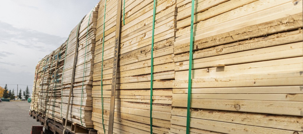 A truck loaded with neatly stacked, light-colored lumber secured by green straps is parked on a broad road under a cloudy sky, suggesting industrial activity.