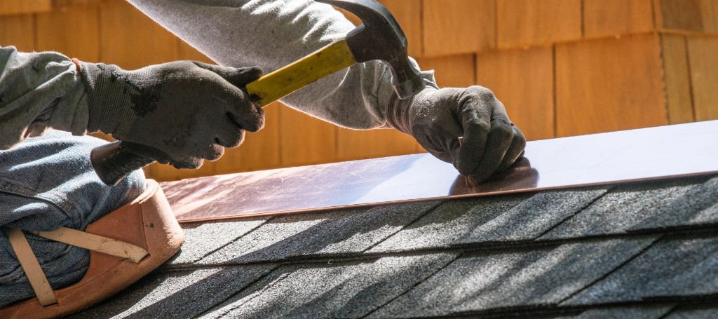 A roofer in work gloves hammers a nail into a shingle on a sunlit wooden roof, focused on careful, hands-on construction.