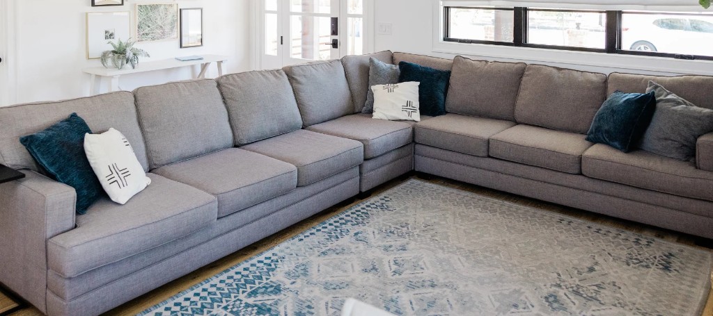 Bright modern living room with a large gray L-shaped sectional, teal and white accent pillows, and a blue patterned rug, creating a calm, inviting feel.