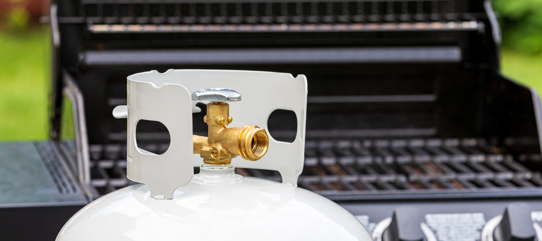Close-up of a white propane tank's brass valve and protective collar in front of an open black gas grill outdoors