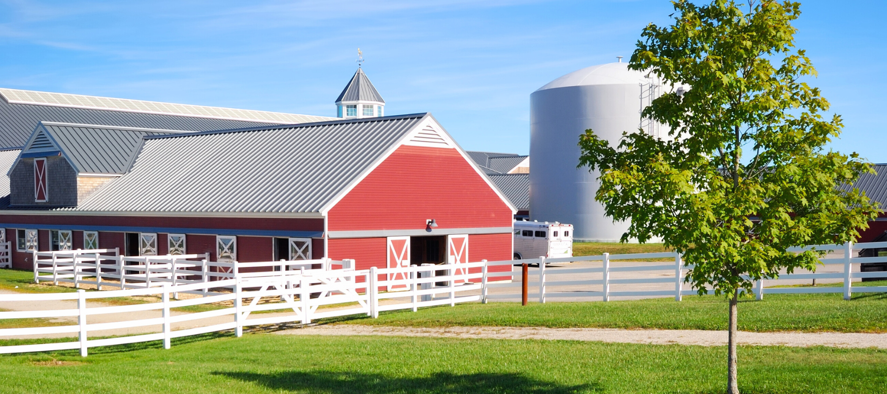 Red barn with white trim and fenced paddocks beside a silver silo beneath blue skies