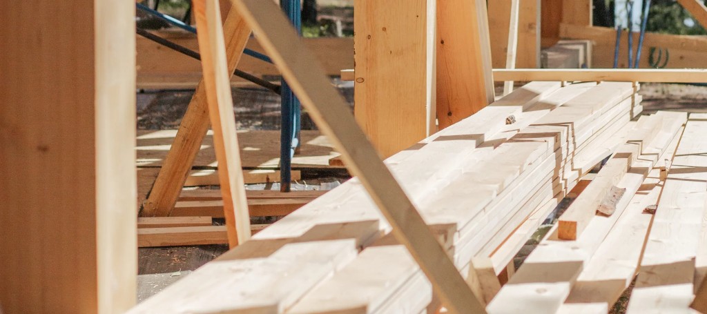 Sunlit timber framing and neatly stacked pale lumber fill an active construction site, conveying careful craftsmanship and steady progress.