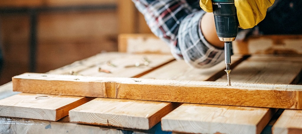 Close-up of a woodworker using a power drill on timber planks in a workshop, conveying hands-on craftsmanship and construction.