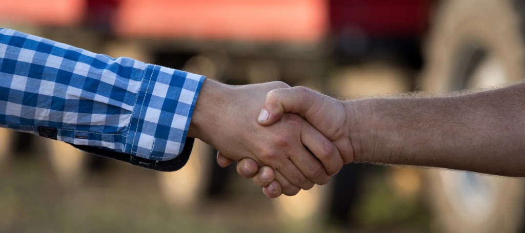 Two people clasp hands in a firm handshake, one wearing a blue checked shirt, suggesting agreement, partnership, or a friendly business meeting.