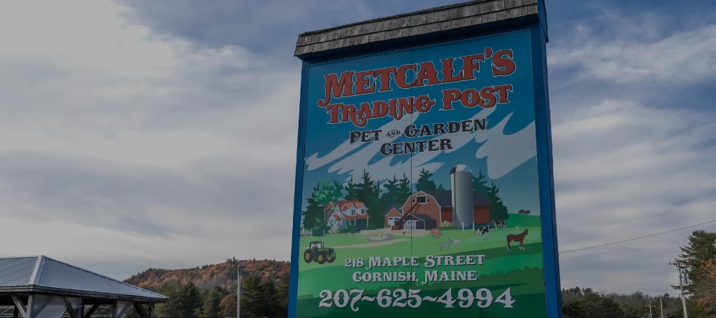 A roadside sign for Metcalf's Trading Post Pet Garden Center in Cornish, Maine, advertises pet supplies and shows a farm scene with animals.