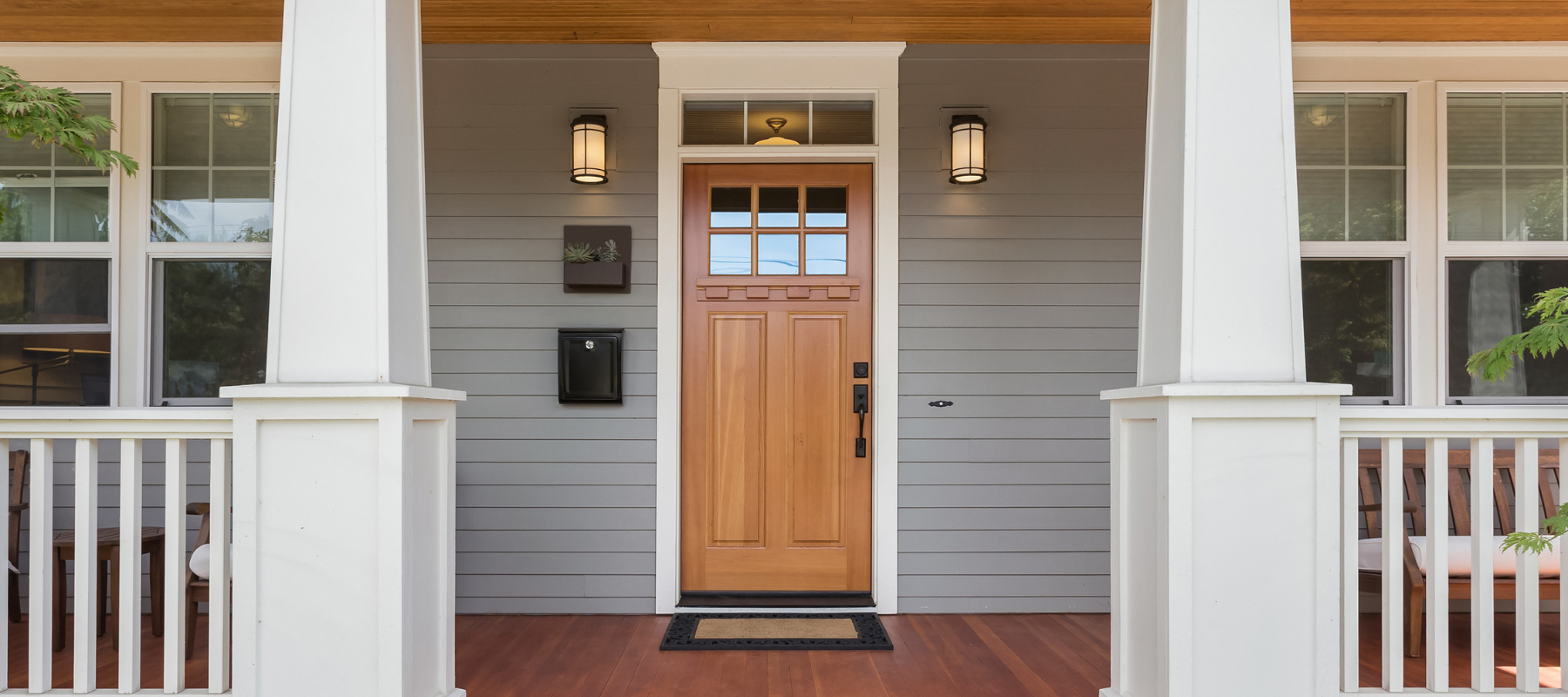 Front porch of a house with a wooden door, gray siding, and black wall lanterns flanking the entry