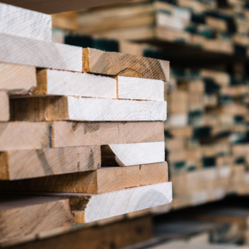 Close-up of stacked unfinished wooden planks in a lumber warehouse, showing rough grain, sawn edges, and blurred timber piles behind.