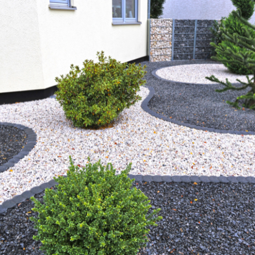 Modern landscaped garden beside a white house, with rounded shrubs, winding dark stone borders, and contrasting white and black gravel.