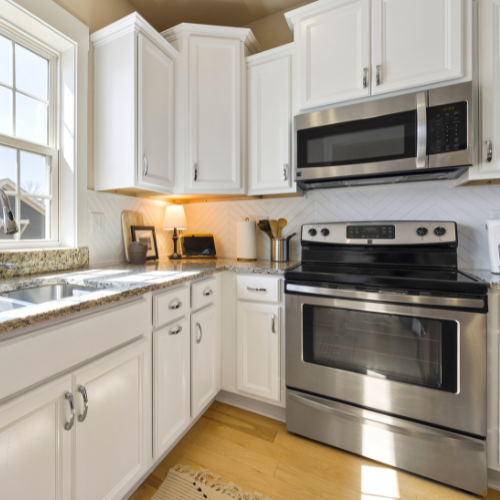 Bright, modern white kitchen with granite counters, stainless-steel microwave and stove, subway-tile backsplash, and warm wood flooring.
