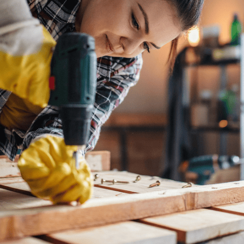A focused worker in yellow gloves drills through a pale wooden board on a workshop bench, with tools and shelves blurred behind him.