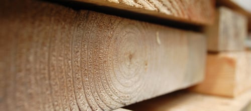 Close-up of stacked wooden planks, highlighting the texture and grain patterns. The image conveys a warm, rustic, and natural tone.