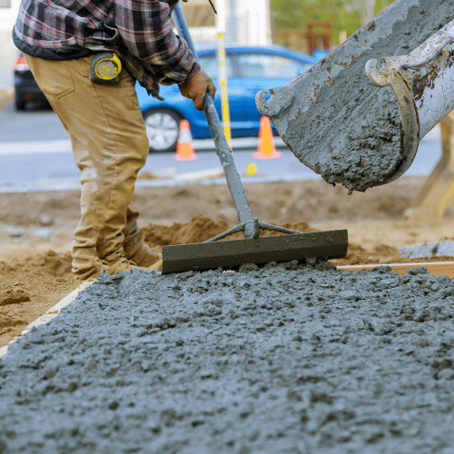A worker in tan pants and plaid shirt levels wet concrete with a tool on a construction site. A blue car and orange cones are visible in the background.