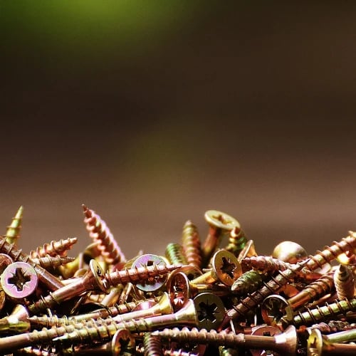 Close-up of a heap of rusty screws and metal fasteners, sharply lit against a blurred dark brown and green background; the cluttered scene feels gritty and industrial.