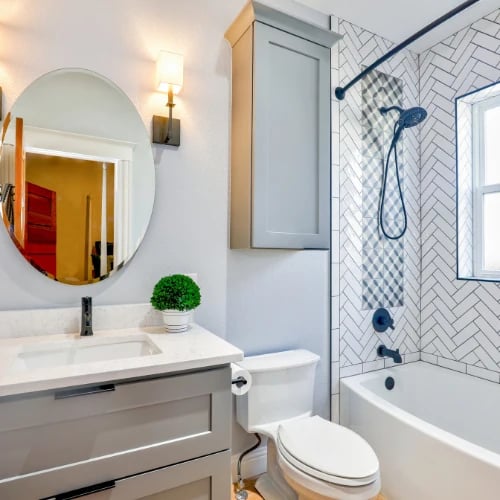 Bright, modern bathroom with a gray vanity and oval mirror, toilet, and white tub with a glass shower screen, dark fixtures, and patterned tile.