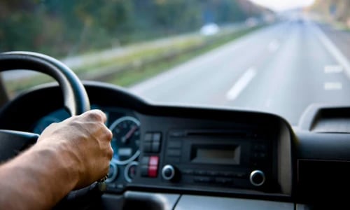 A driver grips the steering wheel inside a car as an open road stretches ahead, conveying a focused, calm journey through a green landscape.
