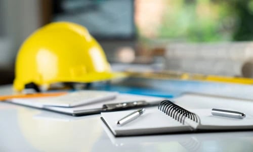 A yellow construction hard hat sits beside blueprints, a spiral notebook, and pens on a table, suggesting careful planning for a building project.