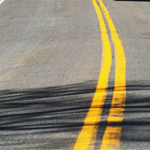 Close-up of a sunlit gray road with curving double yellow center lines, crossed by dark tire marks
