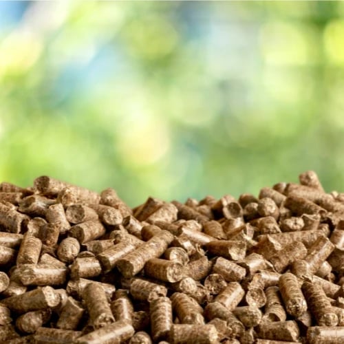 A mound of brown wood fuel pellets fills the foreground, set against a softly blurred green outdoor background, suggesting natural, renewable energy.