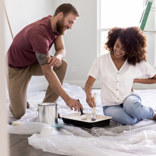 A couple kneels on a plastic-covered floor, smiling as they paint a room with a paint can and tray beside them