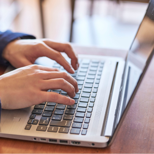 Hands typing on a silver laptop keyboard on a wooden table, conveying focus and concentration. Soft lighting creates a warm and productive atmosphere.